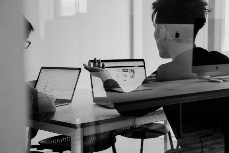 Zwei Personen am Tisch, arbeiten an Laptops und diskutieren. Schwarz-Weiß-Aufnahme.